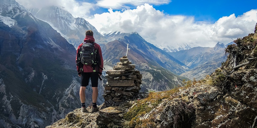 A man stands triumphantly on a mountain peak next to a stack of cairn stones.