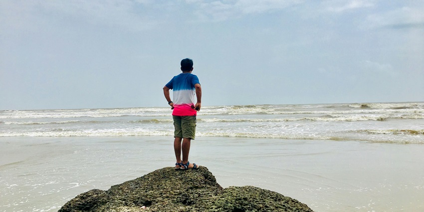 A person wearing a pink and blue t-shirt standing in front of the ocean at Mandrem Beach.
