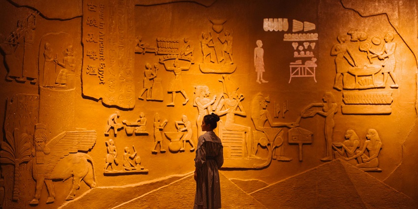 A woman on a backdrop of a large ancient stone structure with carving work done on the wall.