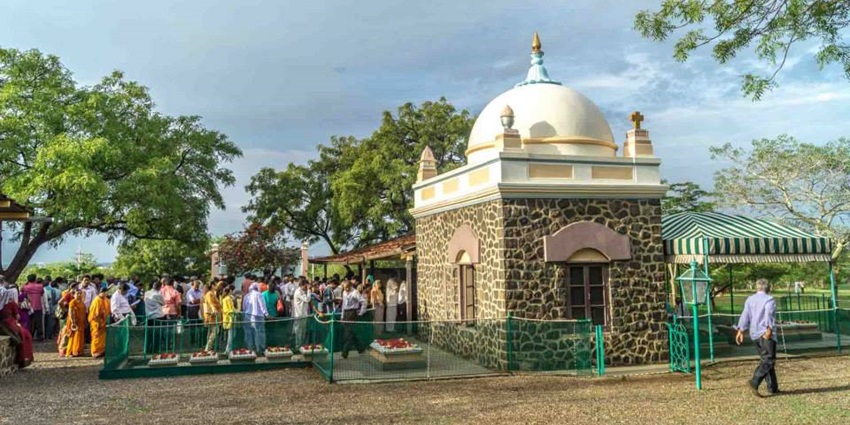 An image of white-domed tomb surrounded by a colourful garden and trees under a clear sky.
