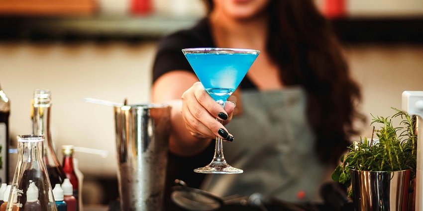 Image of a woman holding glass made a blue drink offering to the people - explore Mia Bella