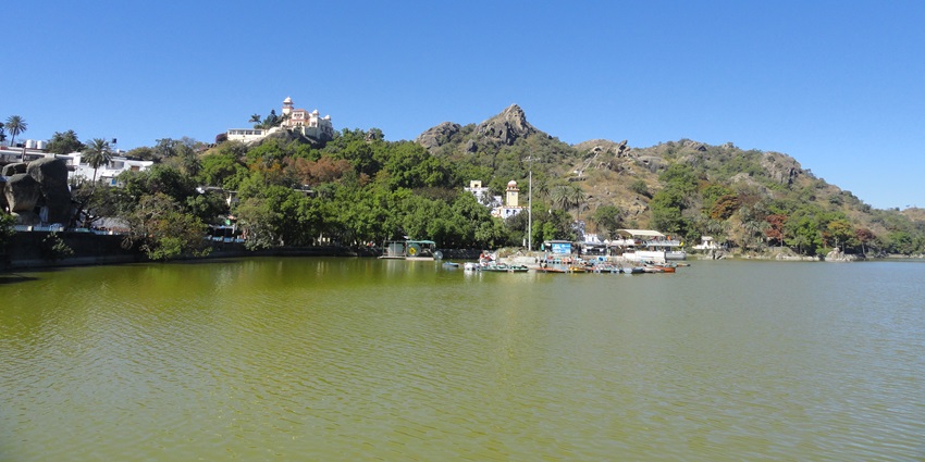 A picture of a lake with clear blue waters in front of a lush green mountain in Rajasthan