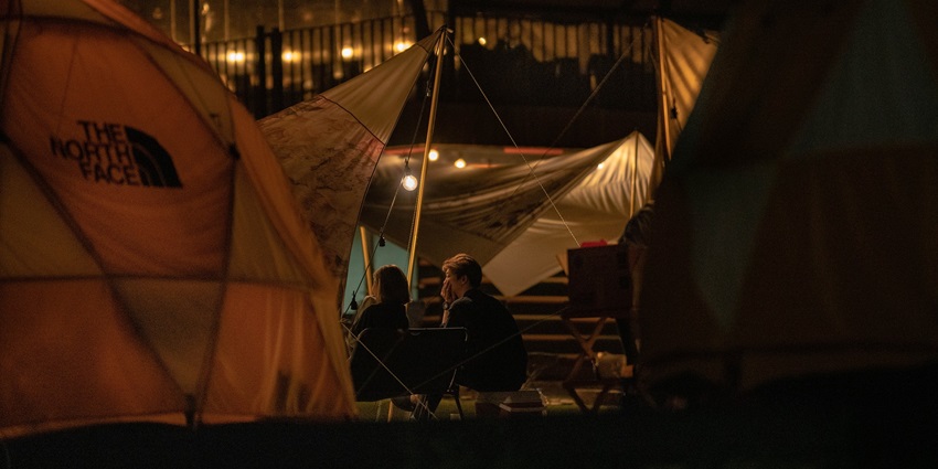Two friends sit outside a tent at night, illuminated by a warm light with trees in the background.