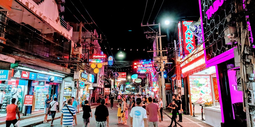 Image of a group of people strolls down a lively street at night, illuminated by city lights.