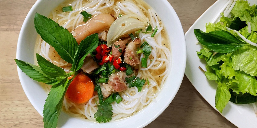 Image of a bowl of freshly cooked noodles garnished with greens and placed on a rustic surface.