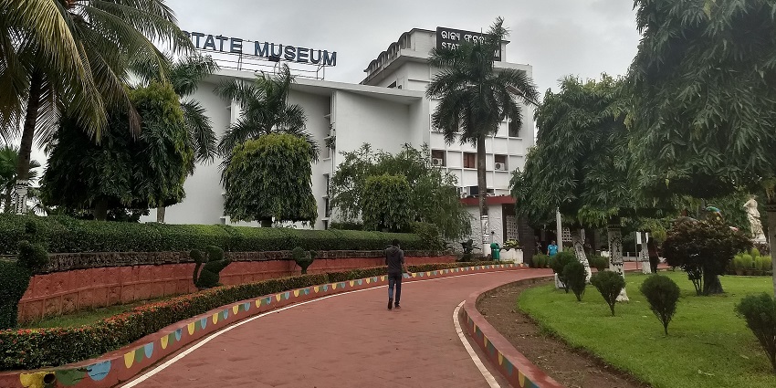 A picture of the Odisha State Museum surrounded by lush green trees and clear skies.