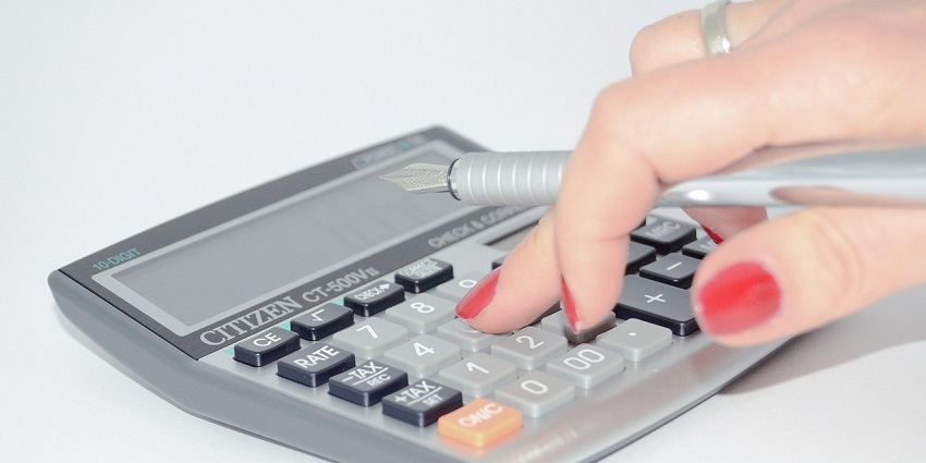 An amazing view of a woman’s hand using a grey calculator kept on a white surface.