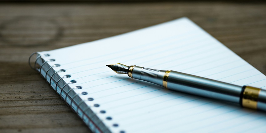 A view of a white open notebook with a golden fountain pen kept on it during the daytime.