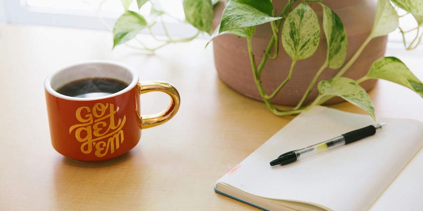 View of an orange cup with black coffee, a notebook, a pen, and a plant lying on a table.
