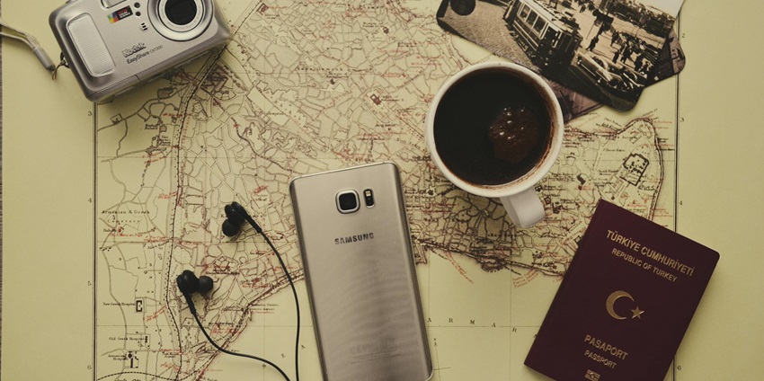 A silver camera beside a black coffee mug, Samsung Galaxy S7, passport, and earbuds.