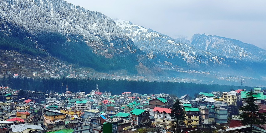 Aerial view of Manali city with buildings nestled among green mountains and forests.