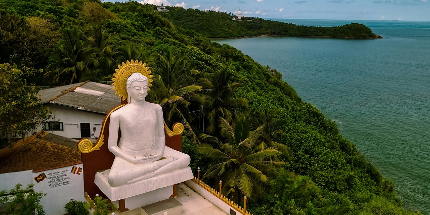 Statue of a person sitting on a bench by the water, with calm waves and a serene setting
