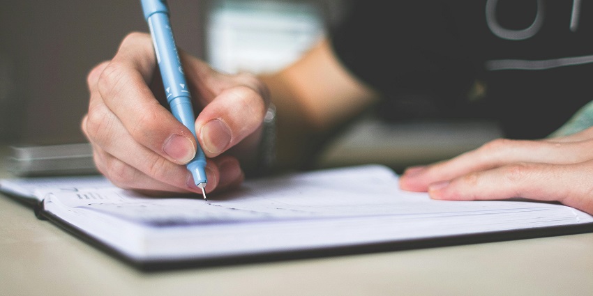A selectively focused view of a person’s hand with a blue pen writing on a notebook.