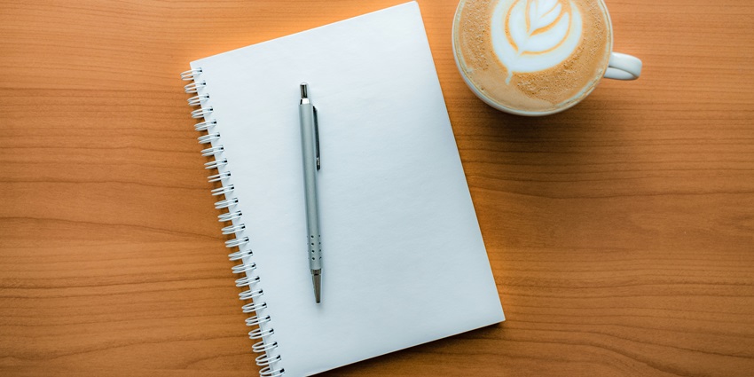 An aerial view of a white open notebook with a grey on top of it with a cup of coffee.