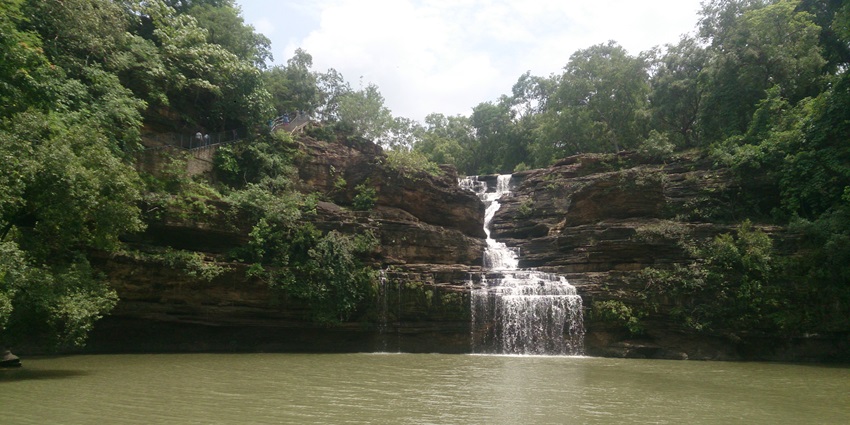 A serene view of Pandav Falls, a waterfall cascading into a clear, emerald-green pool surrounded by dense foliage