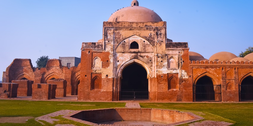 An image of Kabuli bagh masjid front side with one side damaged Tomb wall, blue sky, and green grass.
