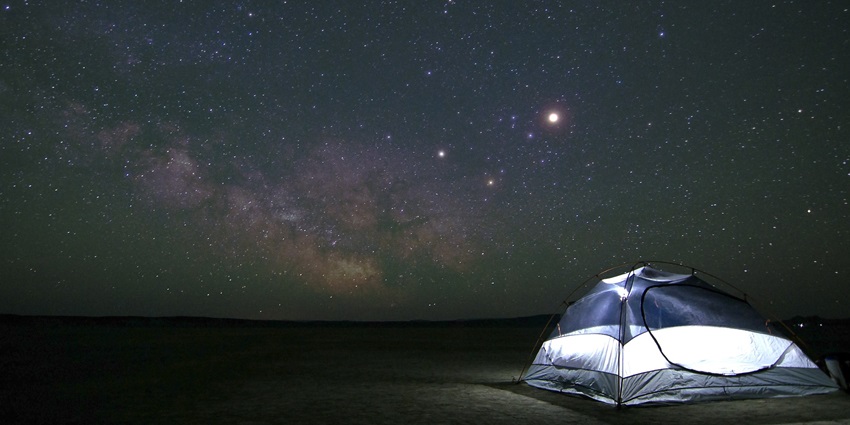 A tent illuminated at night under a clear sky filled with stars and constellations in a remote area.