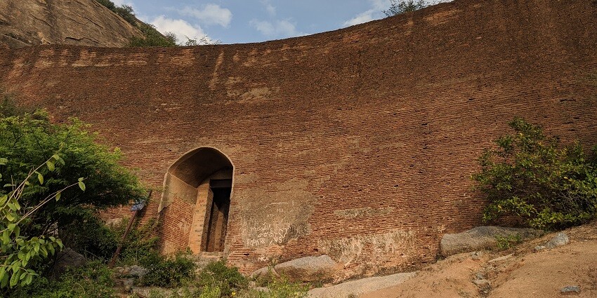 A picture of a tall wall of an ancient fort with an entrance visible in the front besides a few trees