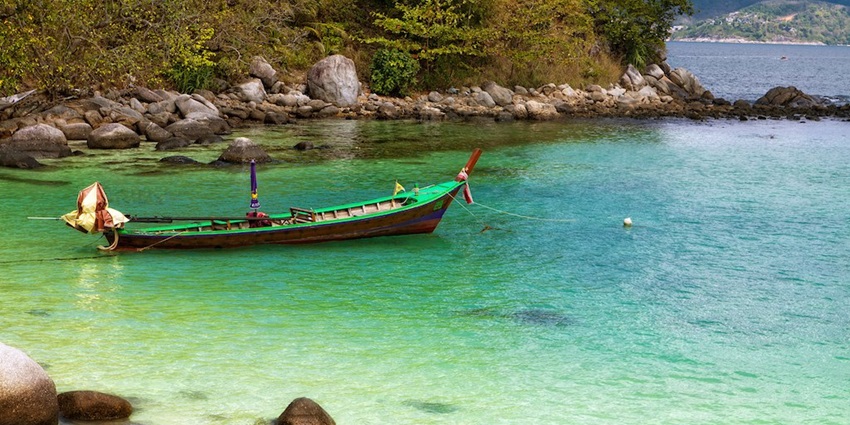 A picture of a boat sitting idle at a shore with the turquoise waters of Phuket in background