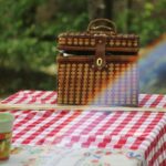 A picnic basket filled with food sits on a wooden table outdoors, inviting a meal