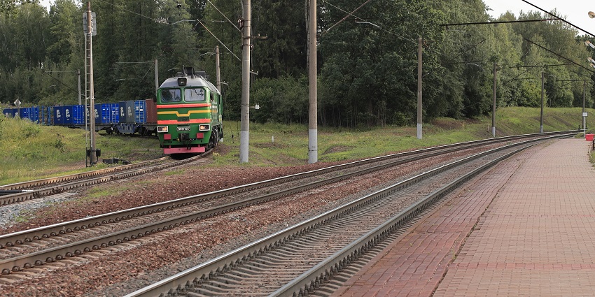 Image of train travelling along tracks next to a lush green forest under a clear sky.