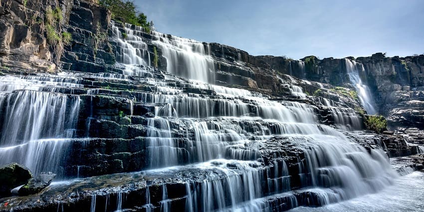 Waterfalls cascading under a clear blue sky, surrounded by lush green landscape and natural beauty.