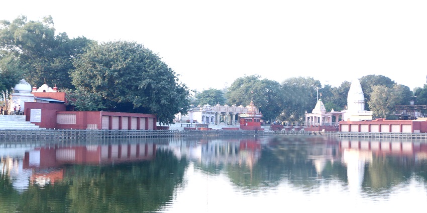 Image of Sannihit Sarovar Lake surrounded by lush green trees and temples during day time