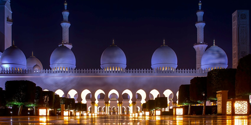 Large white building of a mosque with a decorative plantation and floor in the front.
