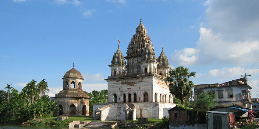 A picture of a temple taken from a distance with a water body in front  of it surrounded by greenery