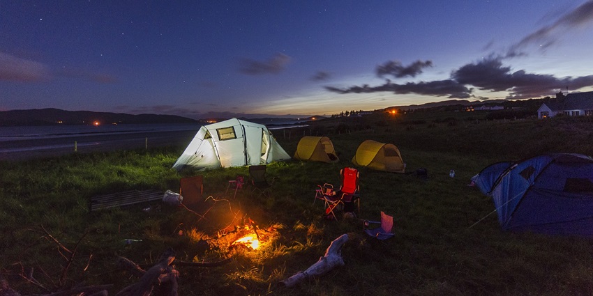 A tent set up near a small campfire in a grassy field, with trees in the background.