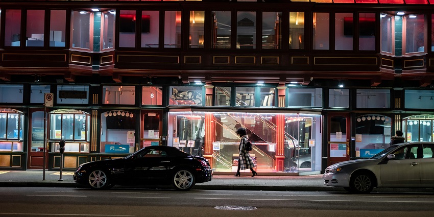 A woman strolls on an urban pavement at twilight, surrounded by city lights.