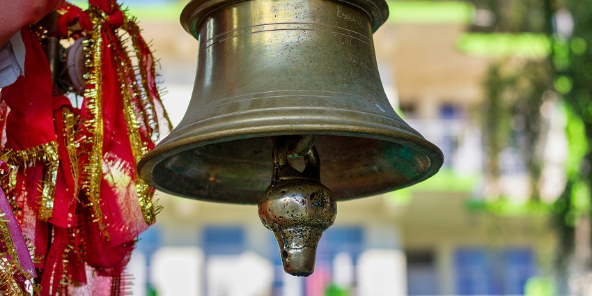 Temple bell hanging from a pole, with intricate details and a clear view of the bell’s clapper.