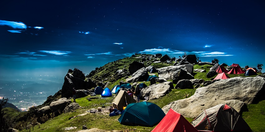 Triund Hill campsite with scattered tents on a grassy slope, surrounded by scenic mountain views