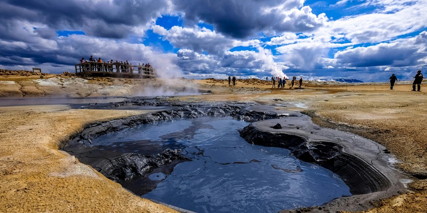 People gather by wooden railings near a steamy hot spring to enjoy on a cloudy day.