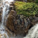 Raging waterfall, similar to Mahadev Paani waterfalls over rocks surrounded by dense green foliage.