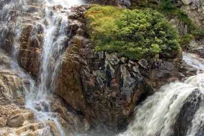 Raging waterfall, similar to Mahadev Paani waterfalls over rocks surrounded by dense green foliage.