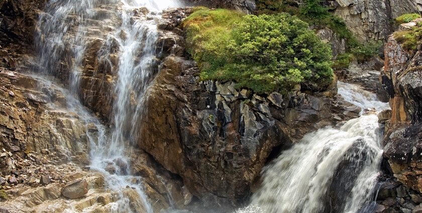 Raging waterfall, similar to Mahadev Paani waterfalls over rocks surrounded by dense green foliage.