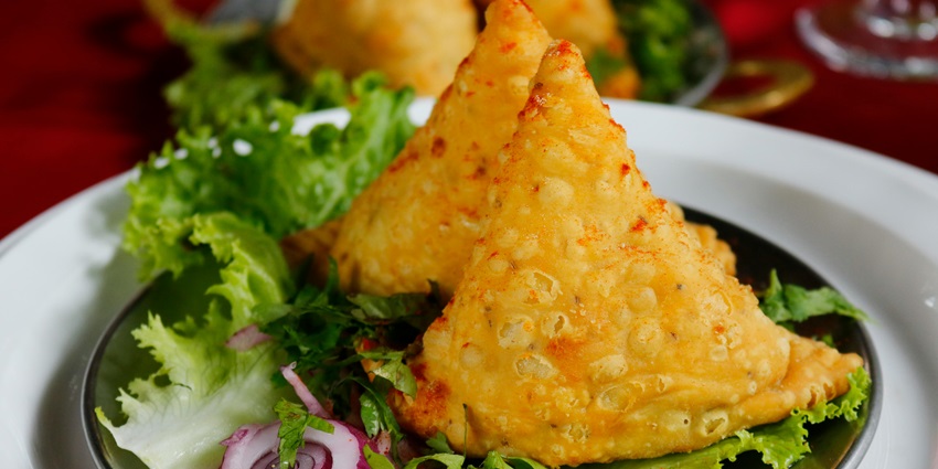 A mouth-watering view of a plate served with 2 fried samosas and green leafs on the side.