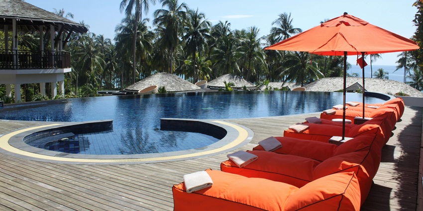 Poolside view of a lodge with seating area, an amazing ambience, and many coconut trees around it.