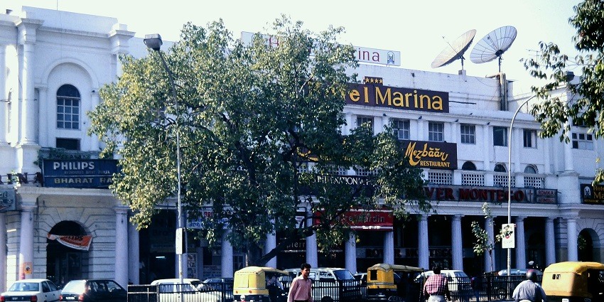 A picture of a hotel in Delhi taken from across the street with vehicles standing in the parking area