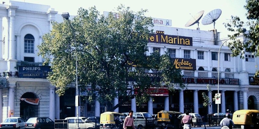 A picture of a hotel in Delhi taken from across the street with vehicles standing in the parking area