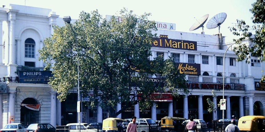 A picture of a hotel in Delhi taken from across the street with vehicles standing in the parking area