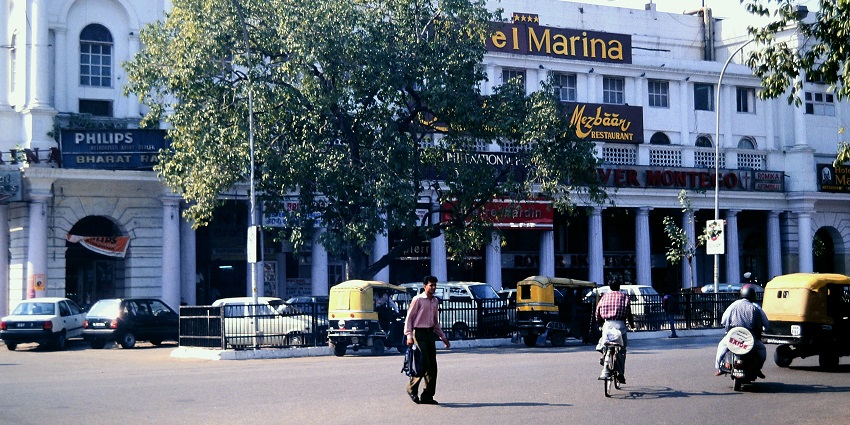 A picture of a hotel in Delhi taken from across the street with vehicles standing in the parking area