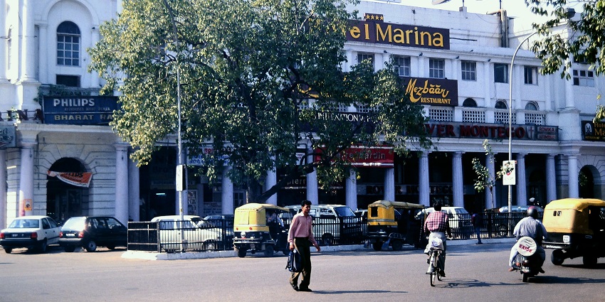 A picture of a hotel in Delhi taken from across the street with vehicles standing in the parking area