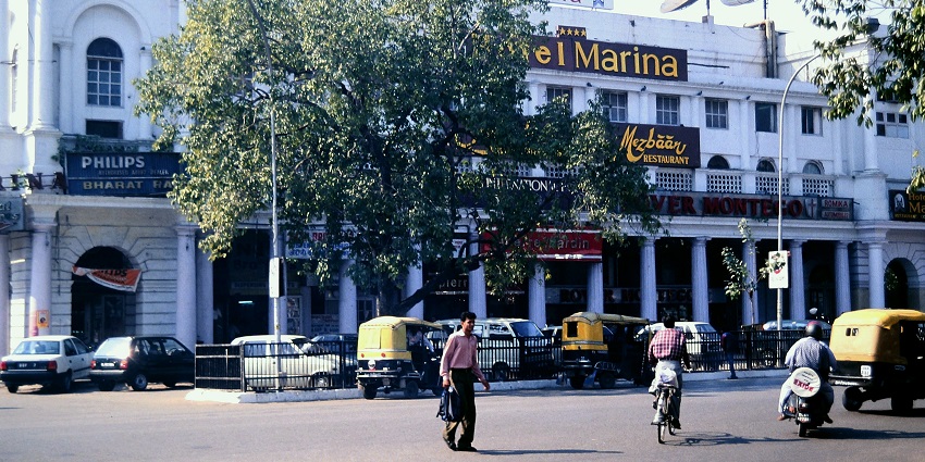 A picture of a hotel in Delhi taken from across the street with vehicles standing in the parking area