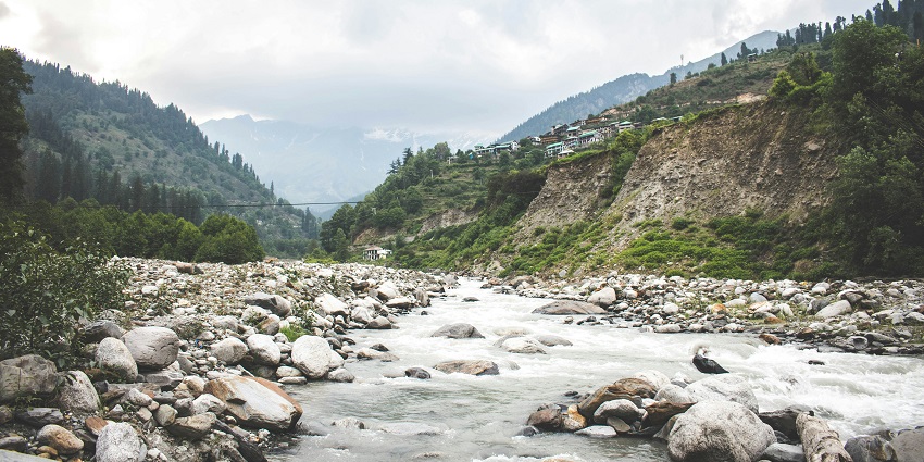 Image of river running through rocks and mountains around with house on the side in Solang Valley
