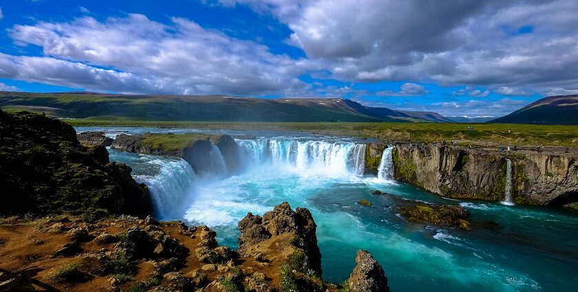 A picture of a serene waterfall with a mountainous view in the background on a cloudy day