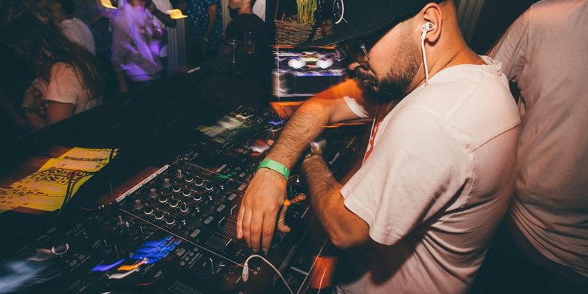 Image of a DJ playing music on his system in a club with full of people dancing on the floor