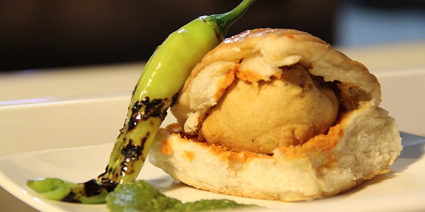 Vada pav served with a spoonful of chutney and a roasted green chilli on a white plate