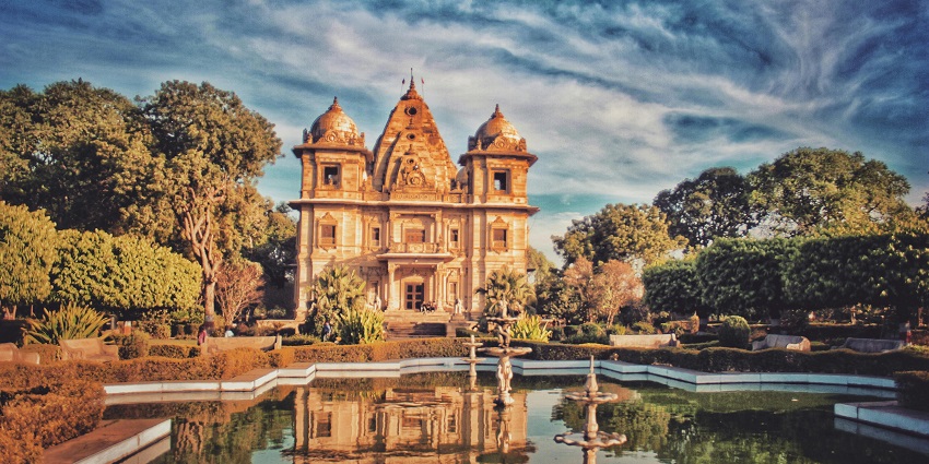 A brown concrete building near a fountain, identified as Balumama Temple, under a cloudy sky.